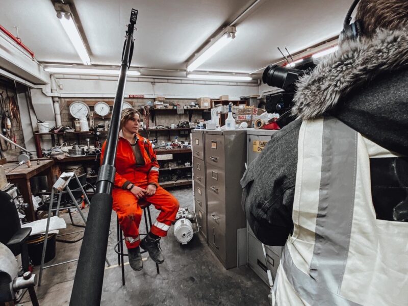 Video production crew member in orange coveralls sits in workshop during behind the scenes filming